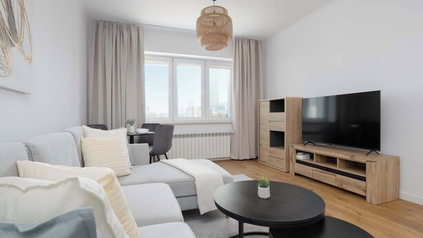 Een moderne woonkamer met een bank, een salontafel, een tv op een houten dressoir en een groot raam met gordijnen.