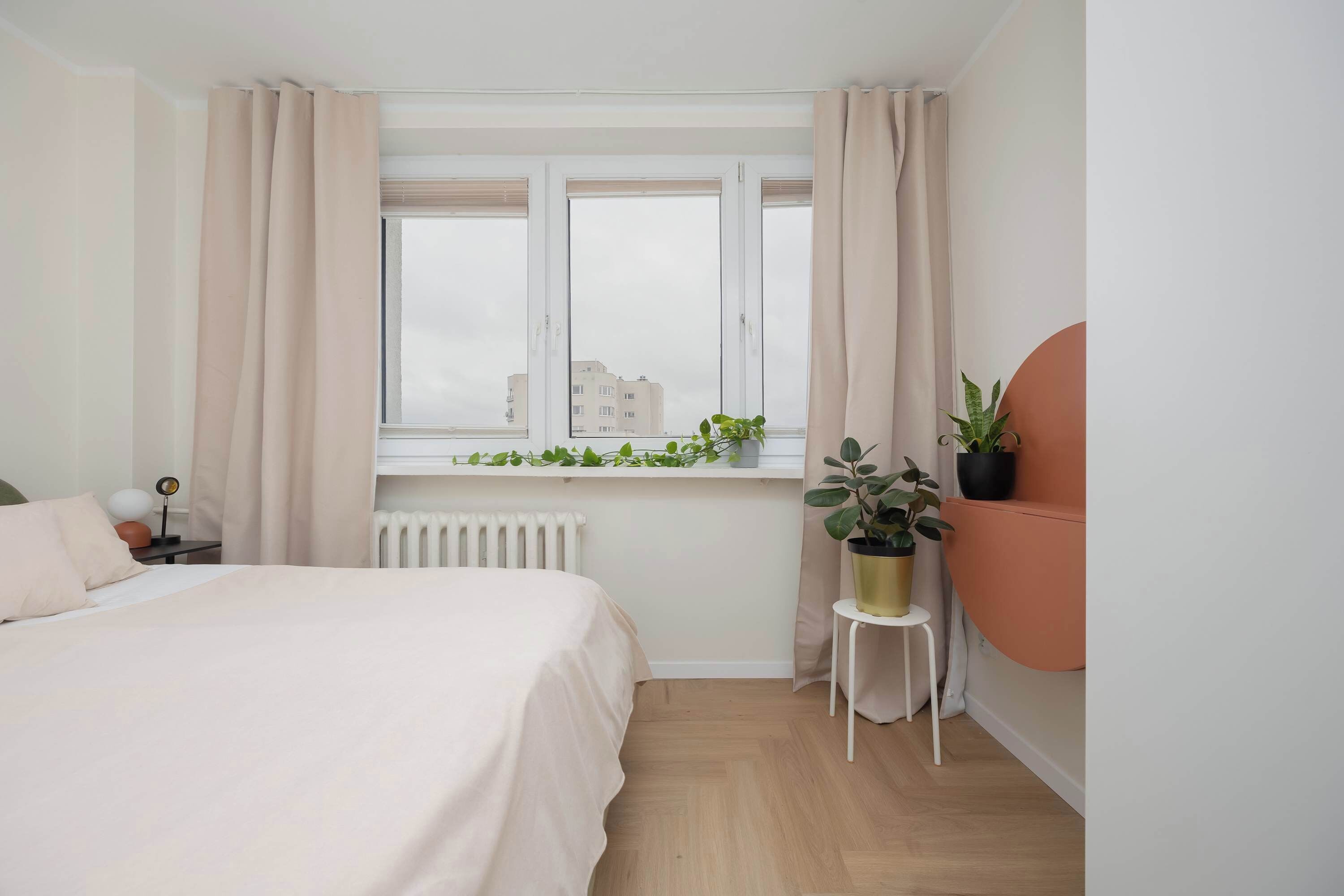 A bedroom with a bed, a window with curtains, a radiator, a small table with a plant, and a wall-mounted circular decoration.