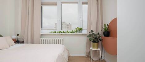 Un dormitorio con cama, ventana con cortinas, radiador, una mesita con una planta y un adorno circular sujeto a la pared.