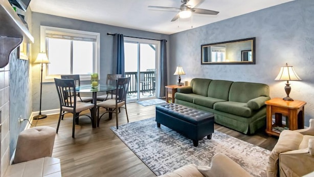 Ein modernes Wohnzimmer mit einem grünen Sofa, einem blauen Hocker, einem Esstisch mit Stühlen und einem Deckenventilator.