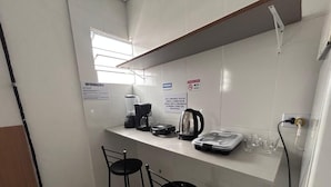 A small kitchenette with a coffee maker, kettle, and a scale. There are two black bar stools and a window above the counter.