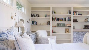 A bedroom with a bed, a chair, a nightstand, a lamp, and a bookshelf filled with books. - Waterfront Saunderstown Estate with Private Dock (North Kingstown)