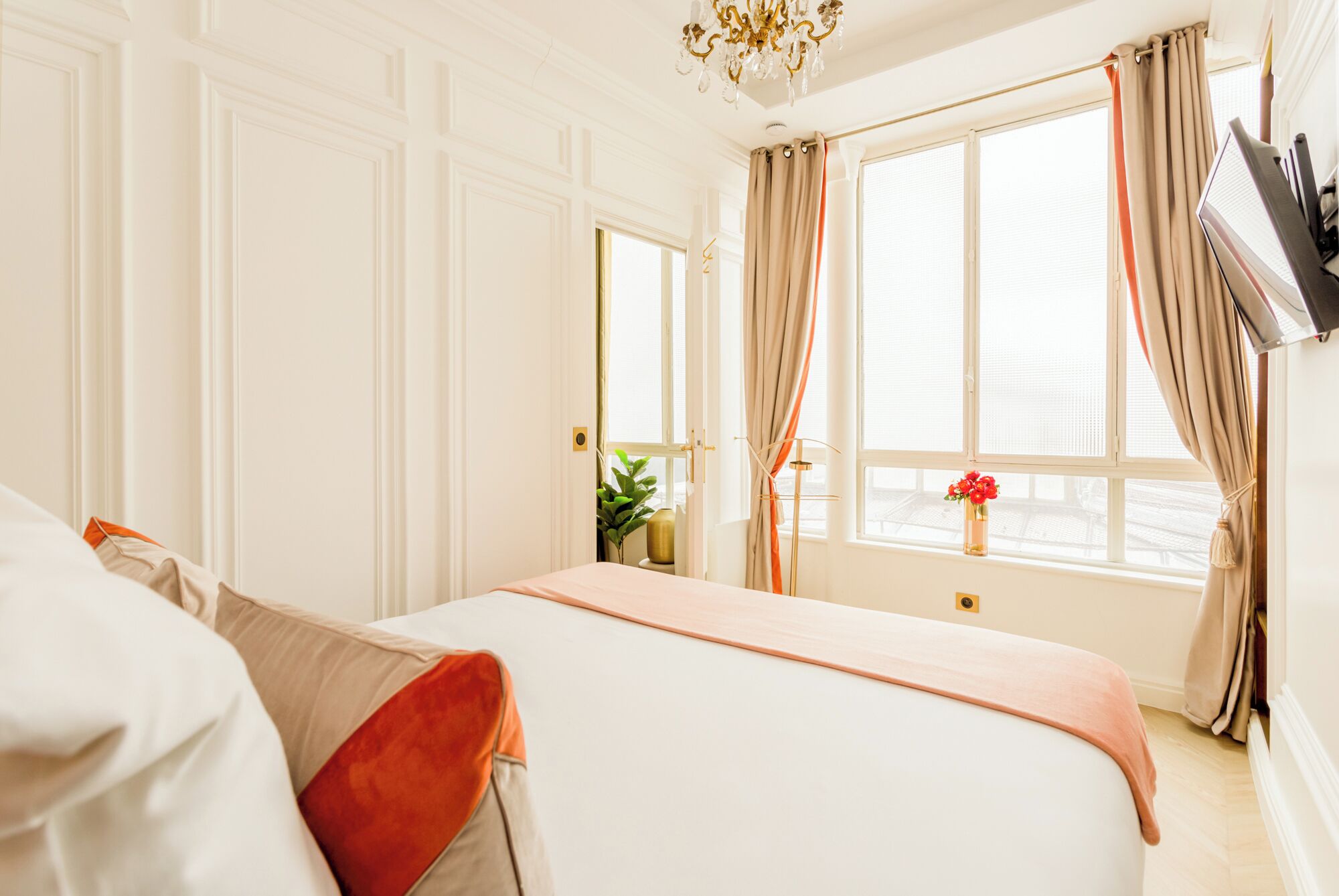 A hotel room with a bed, a window with curtains, a plant on the windowsill, and a chandelier.