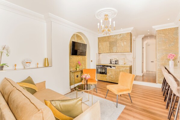 A modern living room with a sofa, a dining area with a table and chairs, a kitchen with a built-in oven, and a hallway leading to another room.