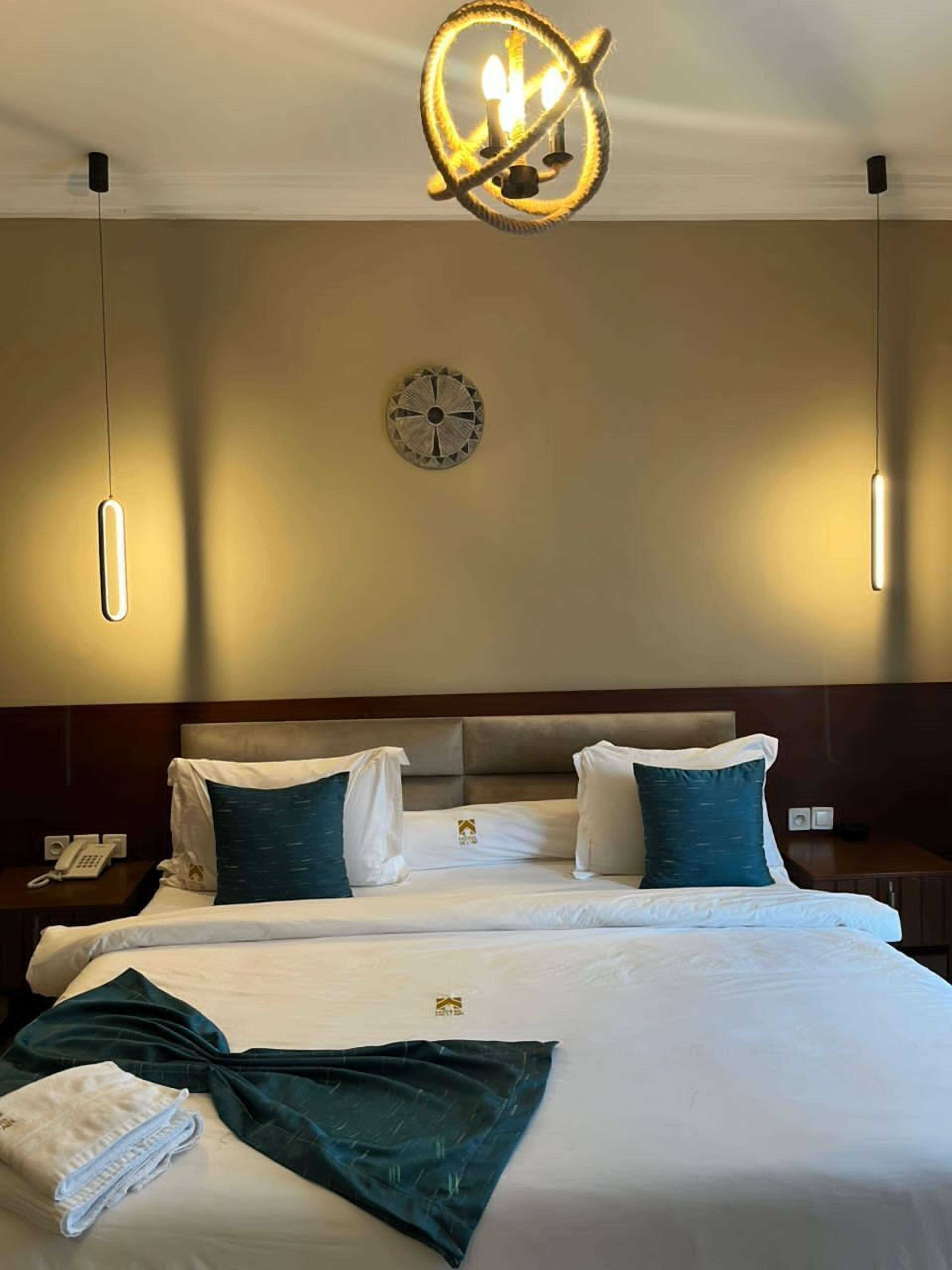 A hotel room with a large bed, two bedside tables, a wall clock, and a modern chandelier.