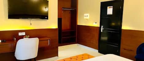 A hotel room with a bed, a desk with a chair, a television mounted on the wall, and a wardrobe.
