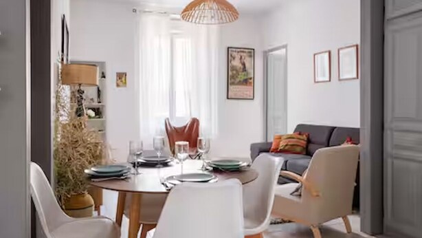 Un coin repas avec une table ronde dressée pour quatre personnes, entourée de chaises blanches. On aperçoit un canapé avec des coussins au fond, et une grande fenêtre ornée de rideaux légers.