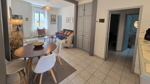 Un salon moderne avec un coin repas, comprenant un canapé, des fauteuils, une table basse et une table à manger. Une grande fenêtre laisse entrer la lumière naturelle, et une porte donne accès à une autre pièce.