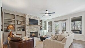 A living room with a stone fireplace, a sofa, a coffee table, and a television mounted on the wall. - Sycamore's Haven in Big Canoe with Hot tub (Big Canoe)
