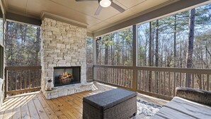 A covered deck with a stone fireplace, wooden flooring, and a view of a forest. - Sycamore's Haven in Big Canoe with Hot tub (Big Canoe)