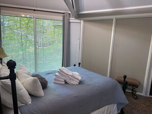 A bedroom with a bed, pillows, a bedside table, a lamp, and a window with blinds. - Treetopper Knoll Chalet in Big Canoe (Jasper)
