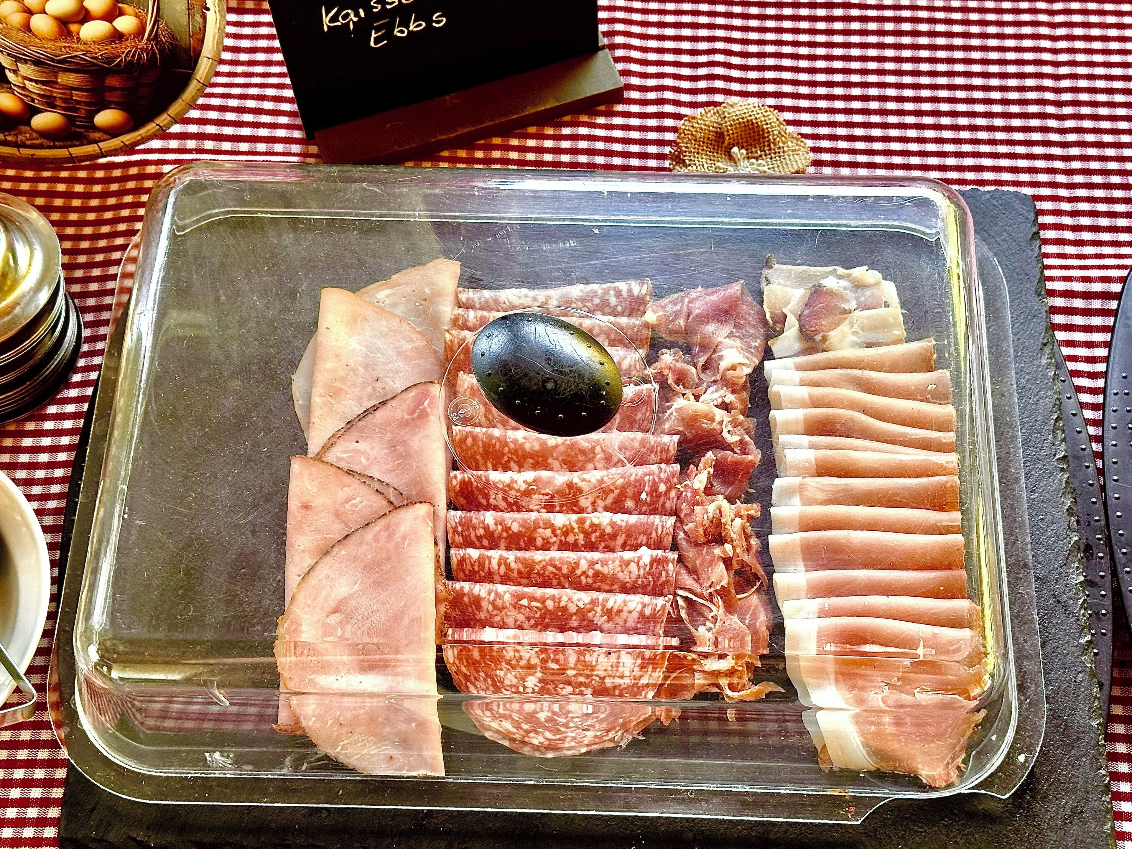 Una vitrina de vidrio con varios tipos de embutidos curados, incluyendo jamón y salami, sobre un mantel a cuadros.