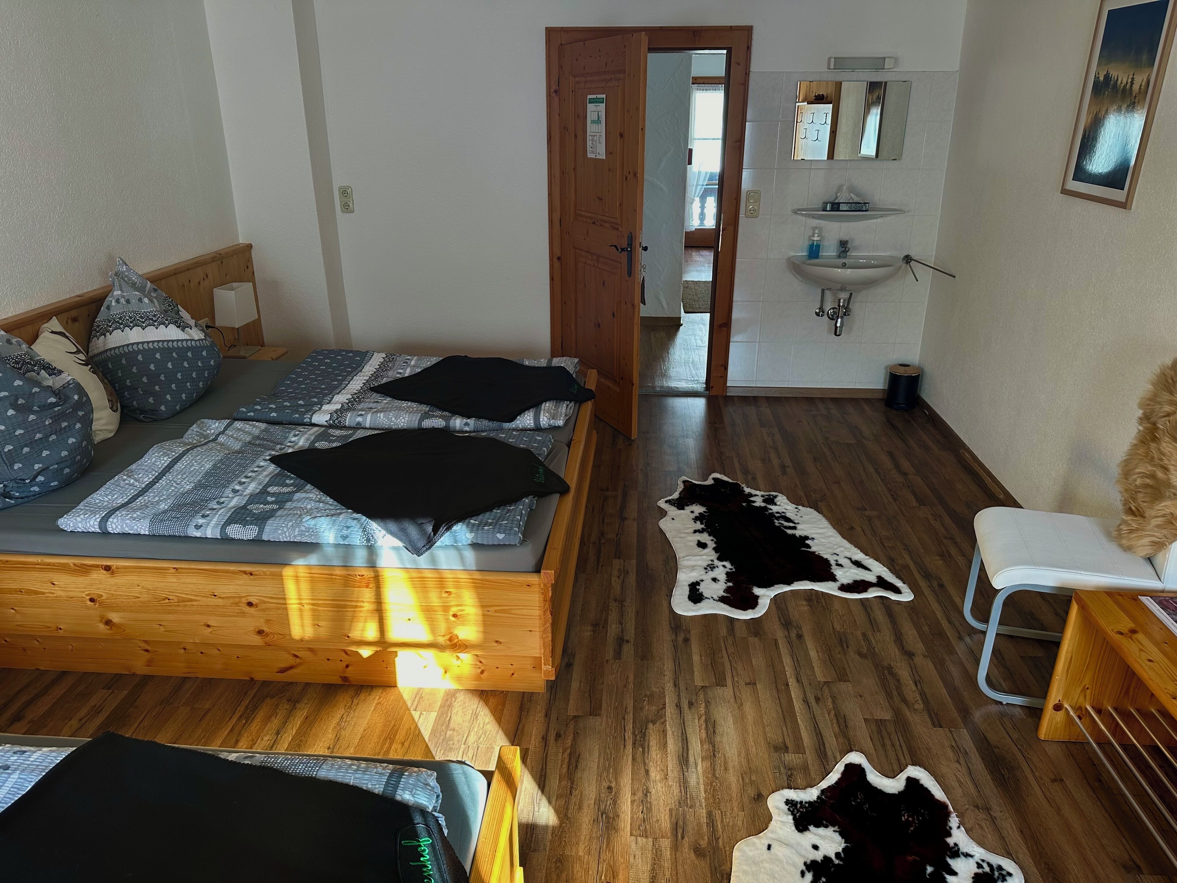 Un dormitorio con una cama de madera, una silla blanca, un tapiz de cuero de vaca y un baño con lavamanos y espejo.