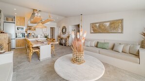 A modern kitchen with wooden cabinets, a dining area with a round table, and a living room with a sofa and decorative plants.