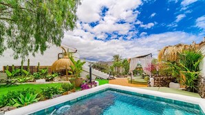 A swimming pool with a stone edge, surrounded by lush greenery, a thatched-roof structure, and a cushioned seating area.