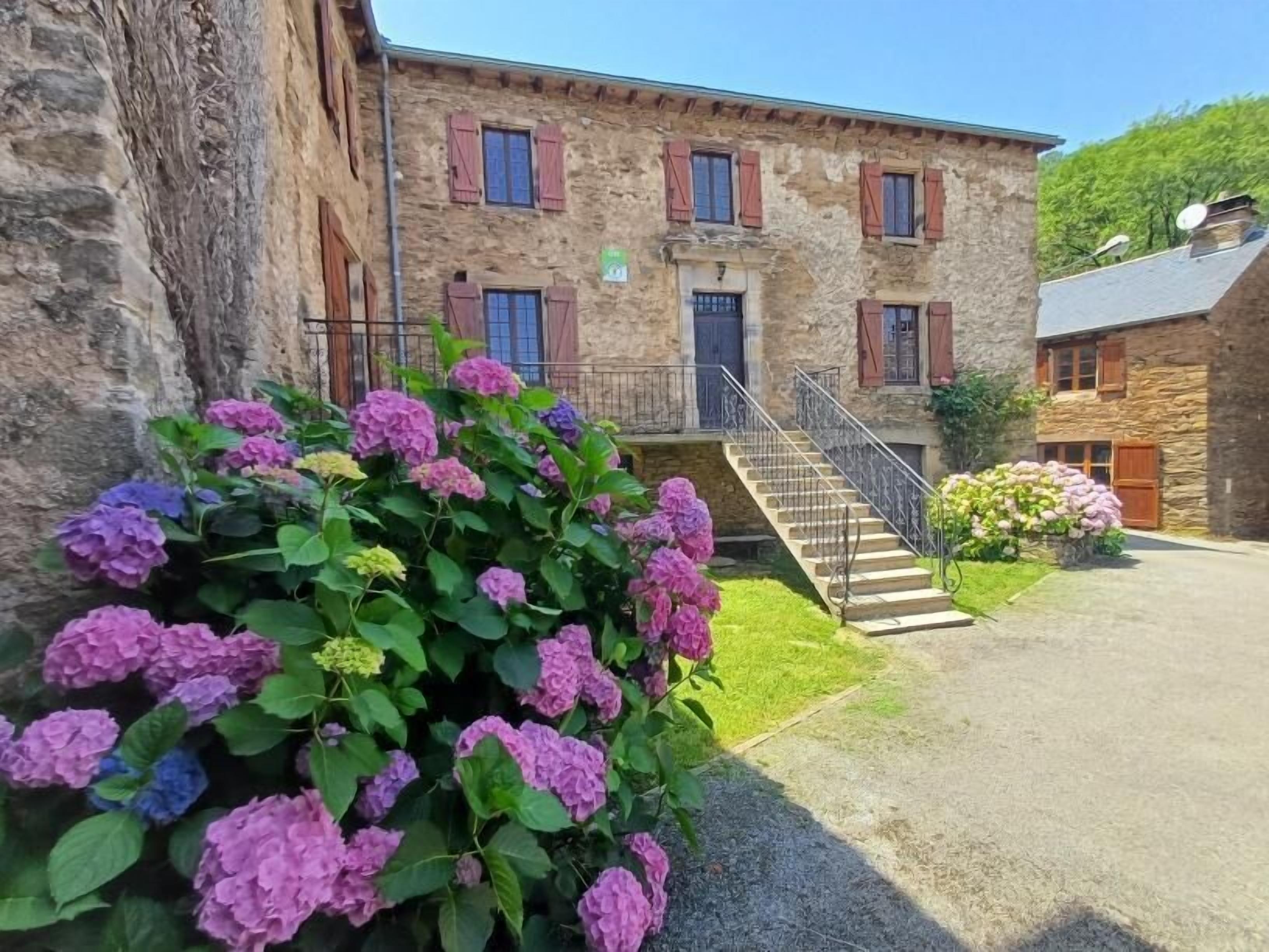 A stone building with a staircase leading to a door, surrounded by greenery and hydrangea bushes.