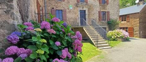 A stone building with a staircase leading to a door, surrounded by greenery and hydrangea bushes.