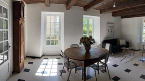 A dining area with a wooden table and chairs, a vase with flowers, and a view of greenery through the windows.