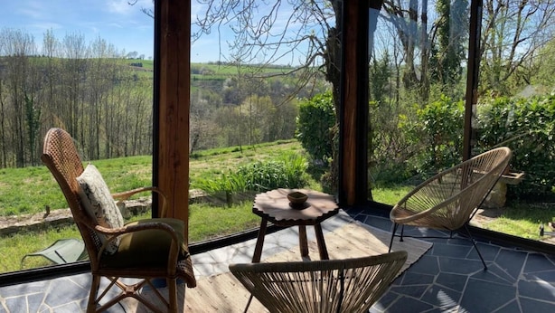 A covered outdoor seating area with wicker chairs and a small table, overlooking a lush garden and distant hills.