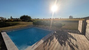 A rectangular swimming pool with a wooden deck surrounding it, set in a landscaped backyard with a clear sky and sunlight.