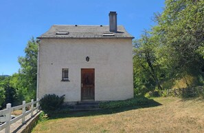 A small, single-story building with a wooden door and a small window, surrounded by greenery and a white fence.
