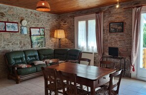 A rustic room with a stone wall, a green leather sofa, a wooden dining table with chairs, a lamp, and a TV on a small table.
