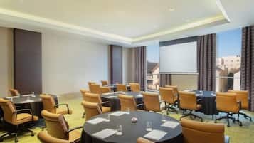 A conference room with multiple tables and chairs, a projector screen, and a view of buildings through the windows.