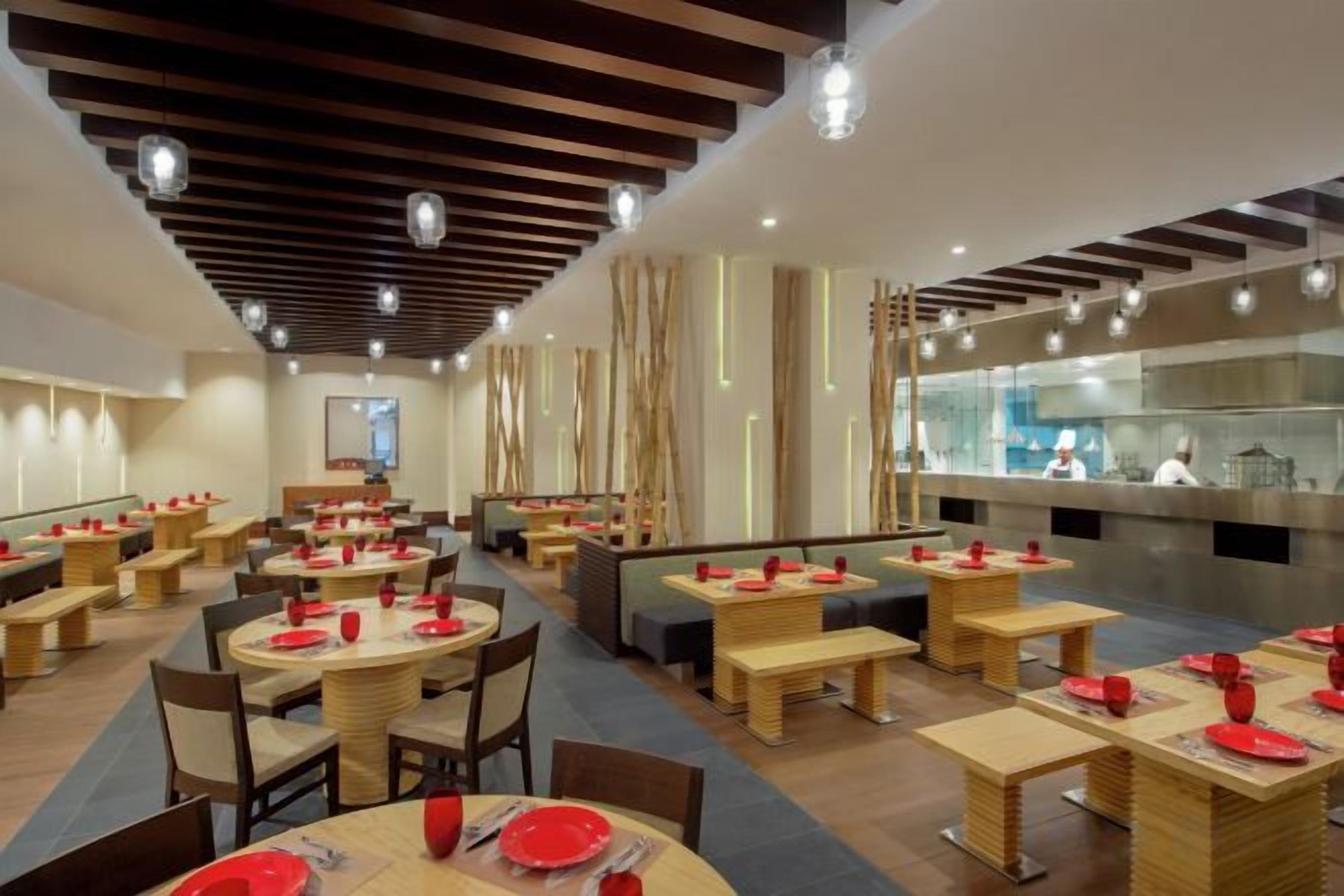 Interior moderno de restaurante com mesas redondas, bancos e uma cozinha à vista.