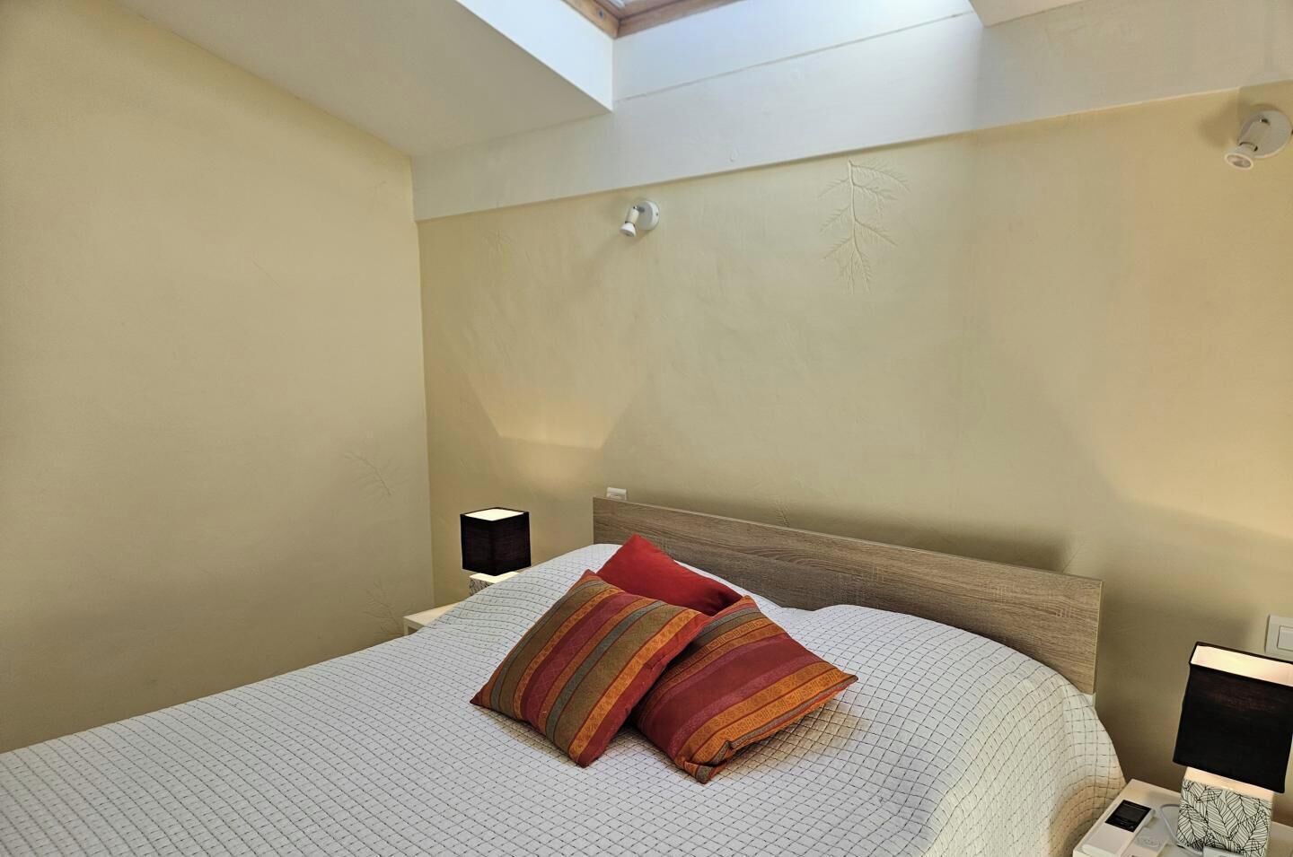 A bedroom with a bed, two bedside lamps, a skylight, and a nightstand with a lamp.