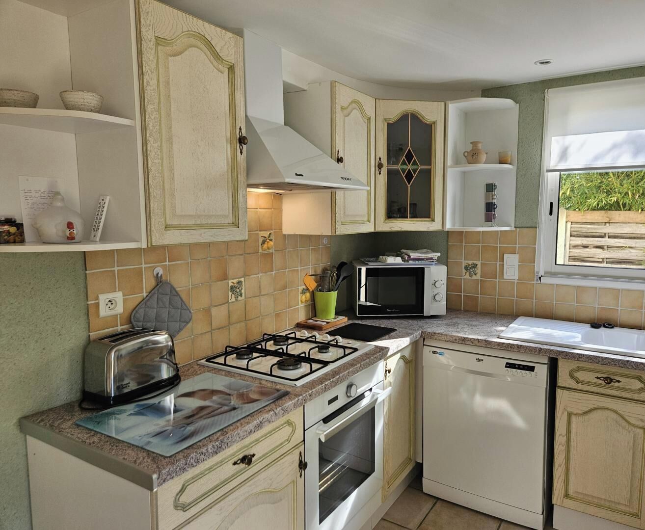 Uma cozinha compacta com fogão a gás, micro-ondas e lava-louças, com armários em tons claros e painel de azulejos.