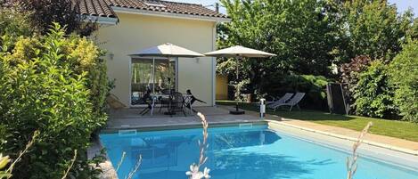 A swimming pool with a patio area, surrounded by greenery and a building with a tiled roof.
