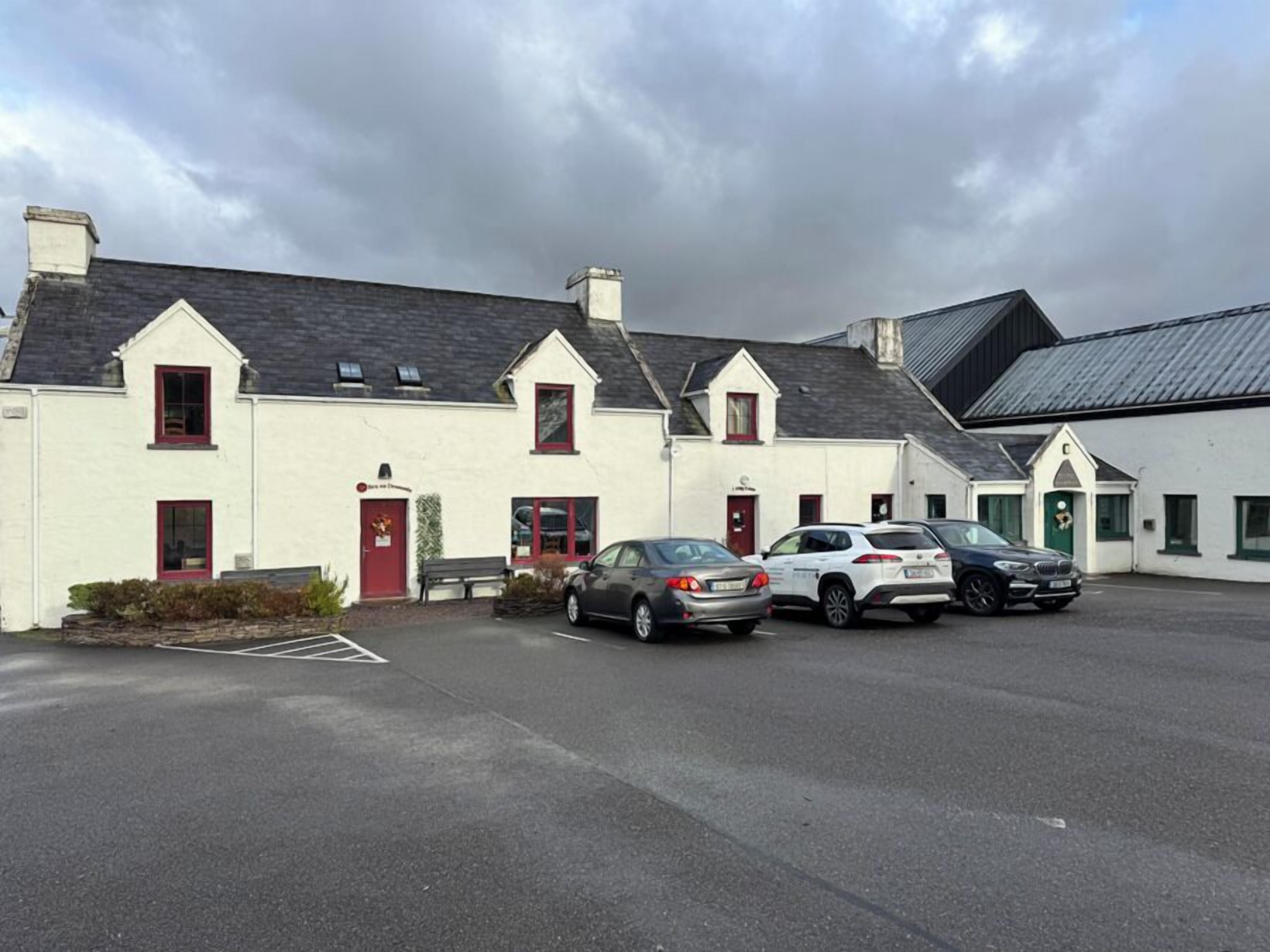 A row of white buildings with red doors and windows, a parking lot with several cars, and a cloudy sky.