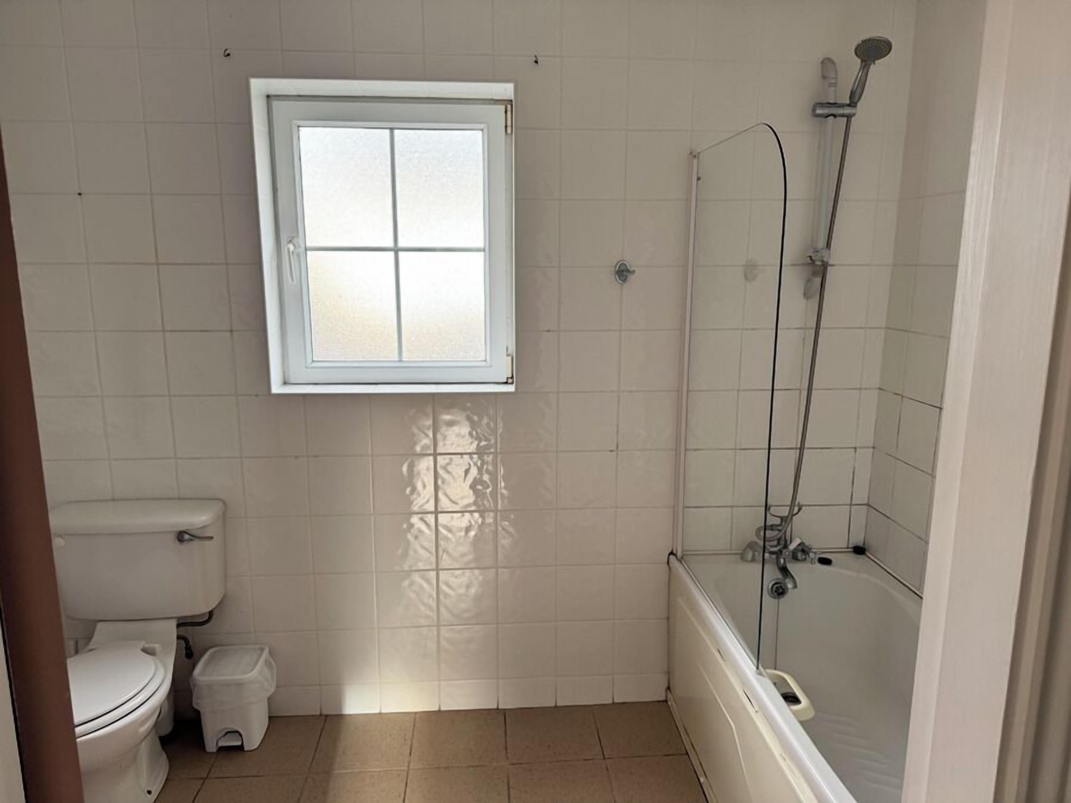 A bathroom with a toilet, a small trash bin, a window, and a shower area.