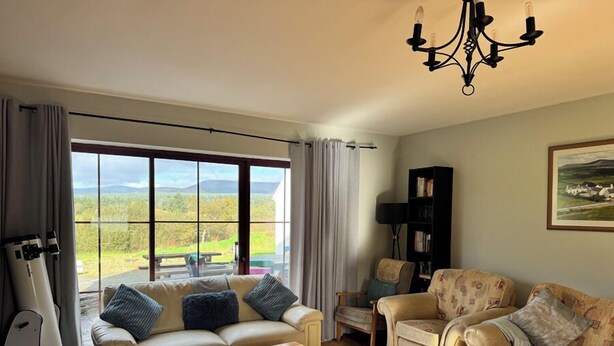 A spacious living room with a large window, a chandelier, and a bookshelf. - Brú na Dromoda Dromid Hostel (Killeenleagh)