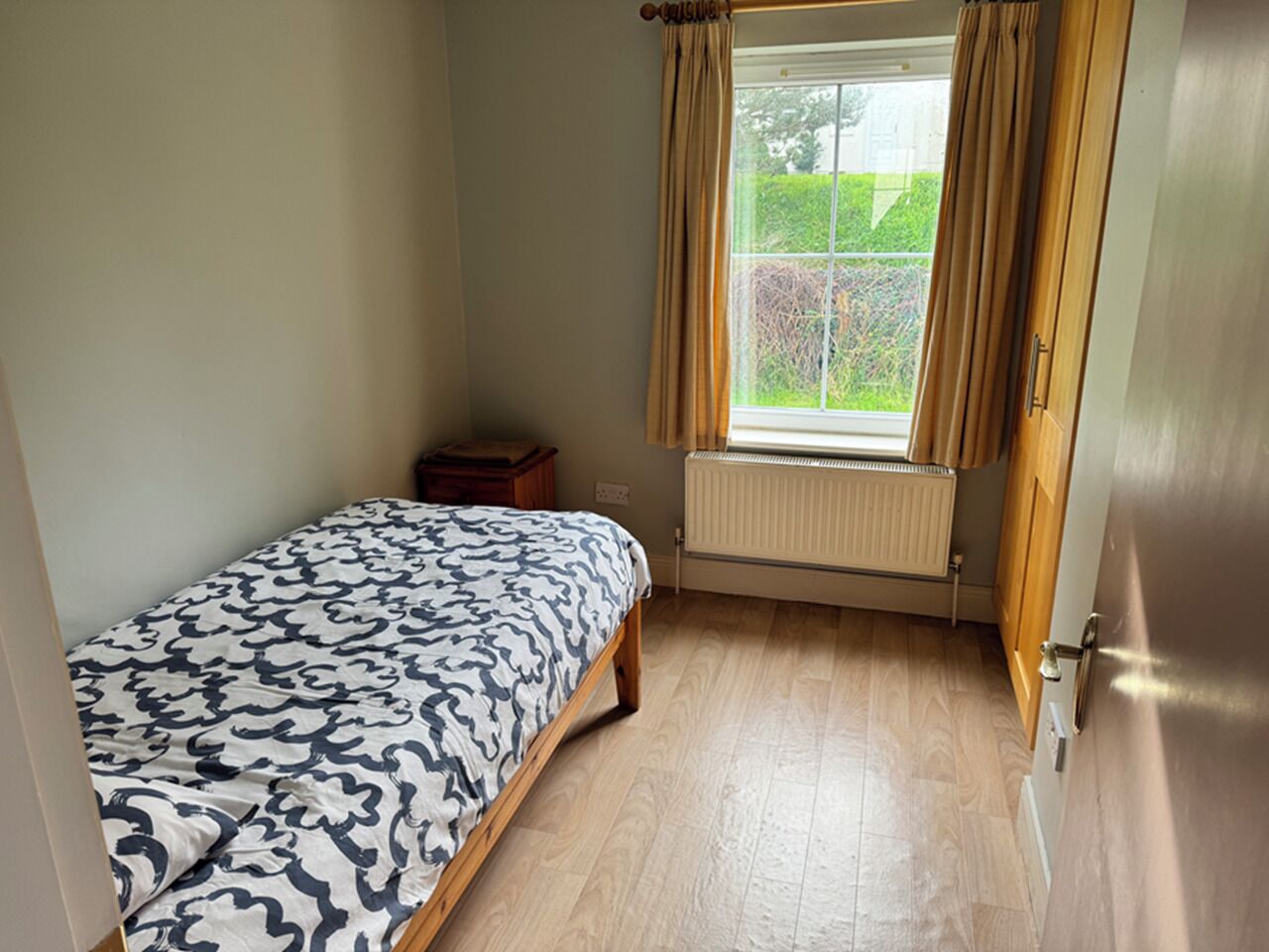 A bedroom with a bed, a radiator, a window with curtains, and a door.