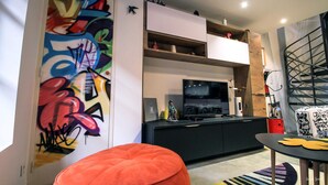 A modern living room with a large colorful graffiti wall, a flat-screen TV, a dining table with a unique yellow table runner, and a staircase with a metal railing.