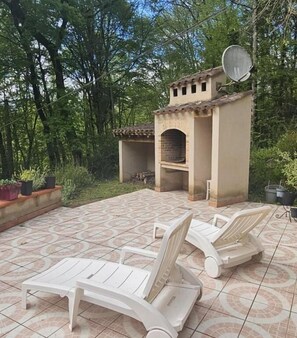 Un patio al aire libre con dos sillones blancos, una zona de barbacoa de ladrillo y plantas en macetas.
