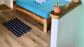 A bedroom with a sloped ceiling, wooden beams, a bed with a blue bedspread, a potted cactus, and a window with a view of greenery.