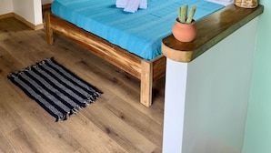 Ein Schlafzimmer mit geneigter Decke, Holzbalken, einem Bett mit blauer Bettdecke, einem Topf mit einem Kaktus und einem Fenster mit Blick auf Grünflächen.