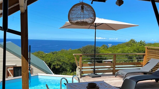 Et poolområde med borde og stole, en pergola med et netbeskyttet parasol og udsigt til havet.