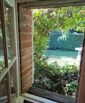 Vista a través de una ventana entreabierta, mostrando un muro de ladrillo, una viga de madera y un seto al fondo.