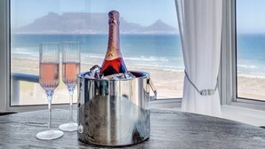 A bottle of champagne in a metal bucket with two glasses on a table by a window overlooking a beach. - Dream beachfront escape with breathtaking Table Mountain, ocean and city views! (Table View)