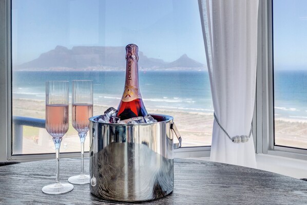 Una botella de champán en un balde de metal con dos copas sobre una mesa junto a una ventana con vista a una playa.