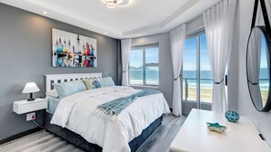 A modern bedroom with a large bed, a bedside table, a nightstand, a window with curtains, and a view of the beach. - Dream beachfront escape with breathtaking Table Mountain, ocean and city views! (Table View)