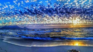 A beach with waves, a sunset, and a mountain in the background. - Dream beachfront escape with breathtaking Table Mountain, ocean and city views! (Table View)