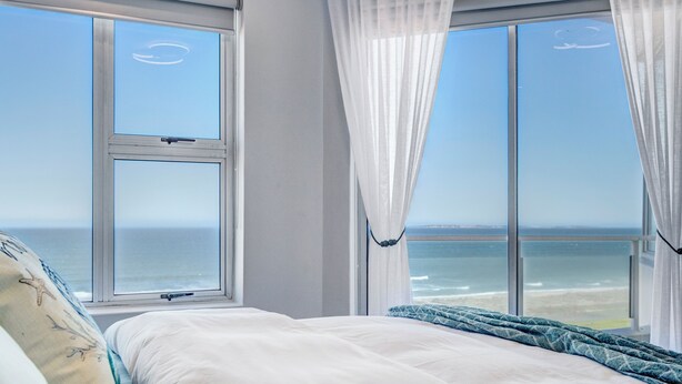 A bedroom with a large window overlooking the ocean, a bed with white bedding, and a view of the beach. - Dream beachfront escape with breathtaking Table Mountain, ocean and city views! (Table View)