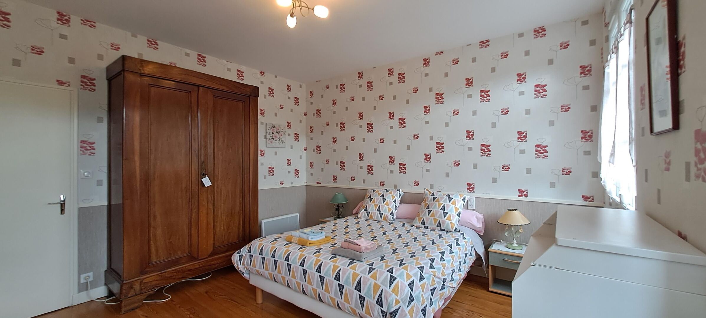 A bedroom with a wooden wardrobe, a patterned bedspread, a bedside table with a lamp, and a window with curtains.