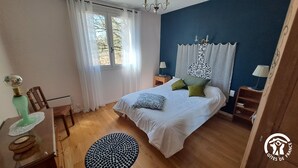 A bedroom with a bed, a nightstand, a lamp, a chair, a window with curtains, and a wooden floor.