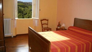A bedroom with a wooden bed, a chair, a nightstand with a lamp, and a view of greenery through the window.