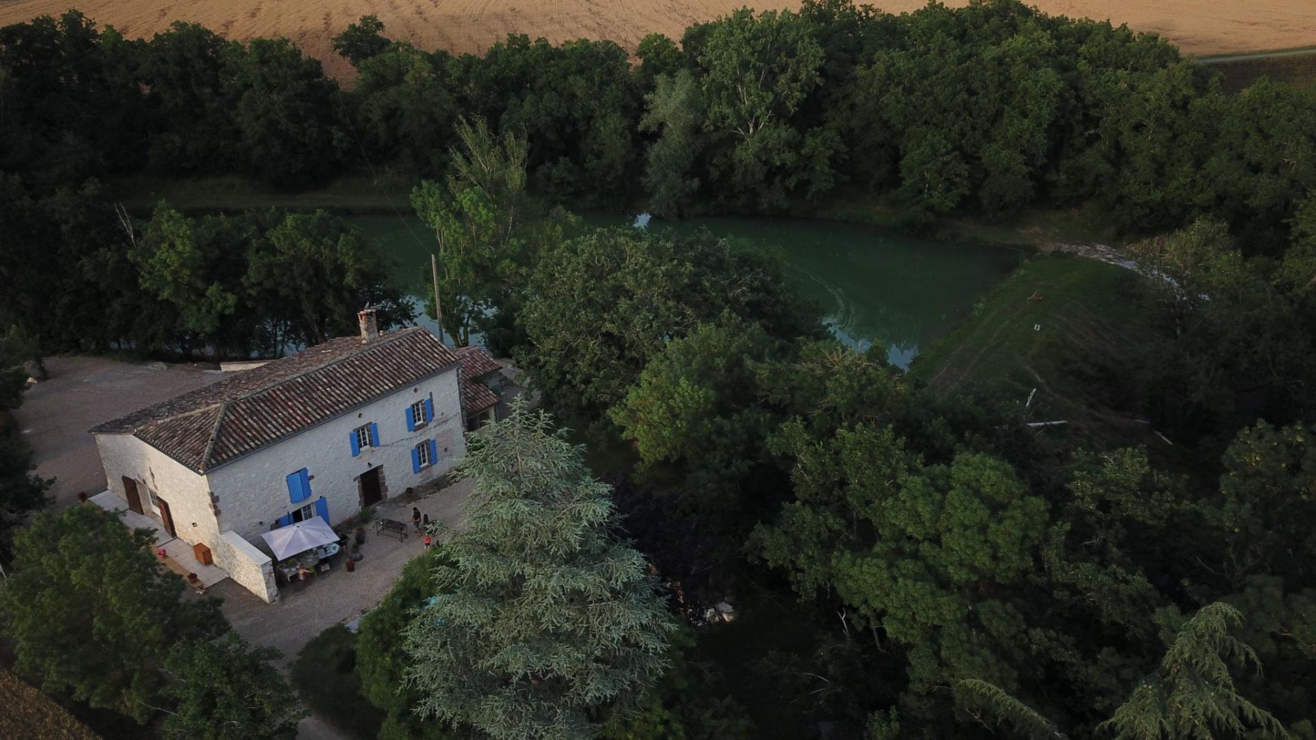 A white building with blue shutters, surrounded by dense greenery and a river.
